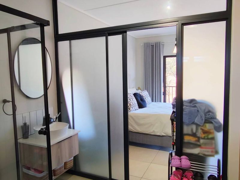 Modern bathroom featuring a glass-enclosed shower and vanity, seamlessly integrated with an adjacent bedroom through a stylish black-framed glass partition.