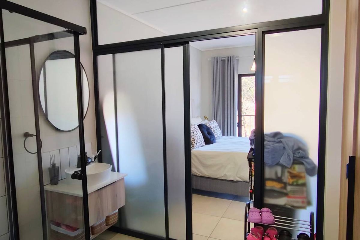 Modern bathroom featuring a glass-enclosed shower and vanity, seamlessly integrated with an adjacent bedroom through a stylish black-framed glass partition.