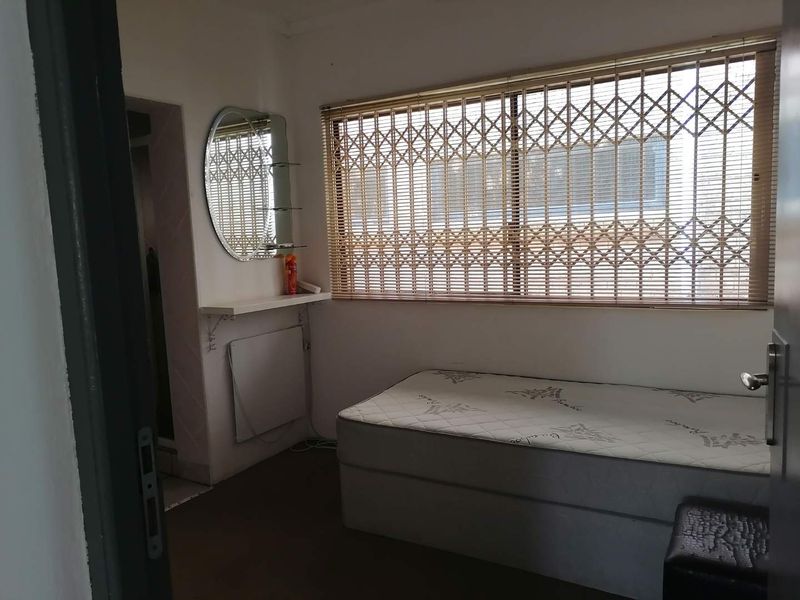 A functional bedroom featuring a window with a security gate, wall-mounted heater, and neutral finishes, offering a practical space for rest.