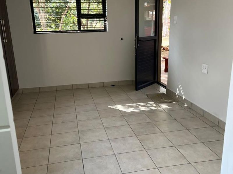 A bright, empty bedroom featuring neutral tiled flooring, a built-in cupboard, and direct access to the exterior through a glass-panelled door.