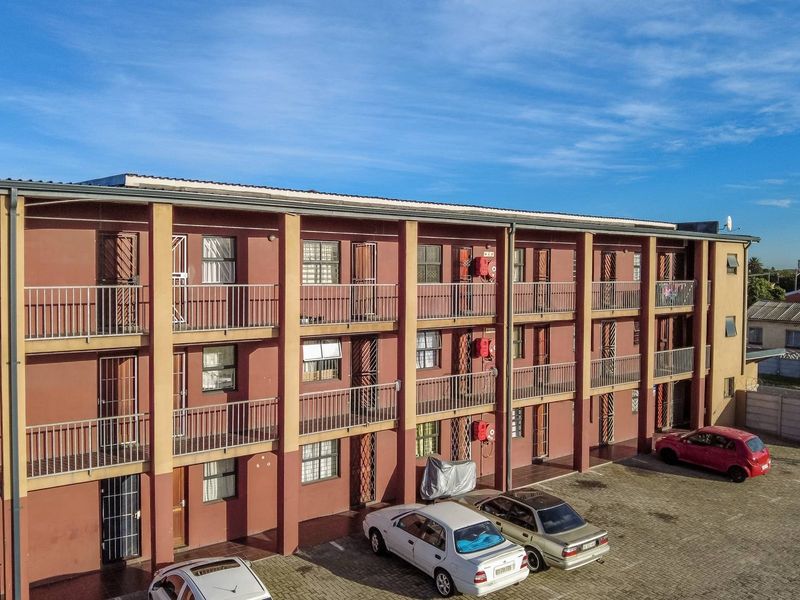 An aerial view of a multi-story apartment complex featuring external walkways, metal balcony railings, and a paved ground-level parking area.