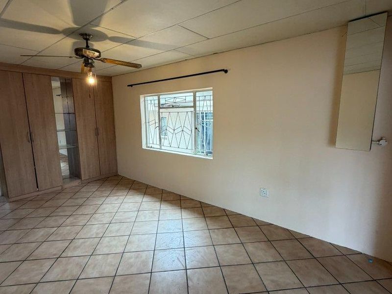 Spacious bedroom featuring tiled flooring, built-in cupboards, a ceiling fan, and security bars on the window.