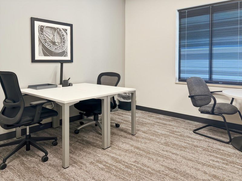 A clean, professional office space featuring neutral walls, carpeted flooring, and a window with fitted venetian blinds.