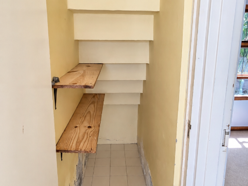 A small under-stair utility storage space featuring tiled flooring and built-in shelving, currently requiring cosmetic wall repairs.