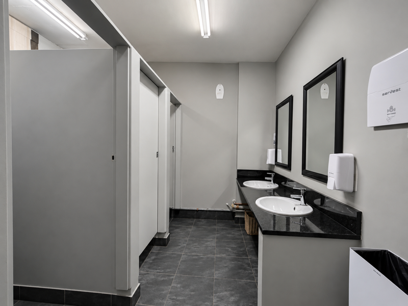 A clean, modern office bathroom featuring a dual vanity with granite countertops, under-mount sinks, and partitioned toilet cubicles with dark tiled flooring.