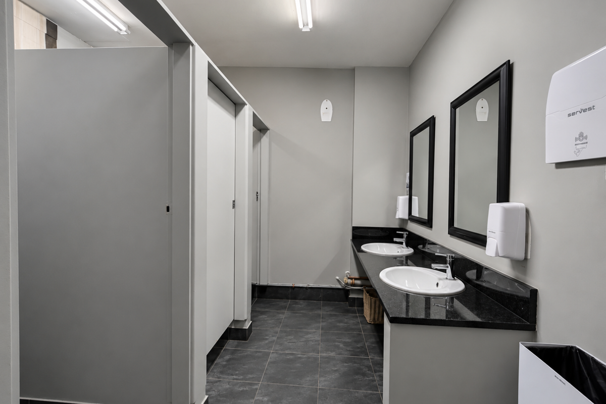A clean, modern office bathroom featuring a dual vanity with granite countertops, under-mount sinks, and partitioned toilet cubicles with dark tiled flooring.