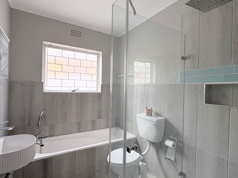 Modern bathroom featuring a built-in bathtub, glass-enclosed shower with a rain shower head, wall-mounted basin, and neutral grey tiling throughout.