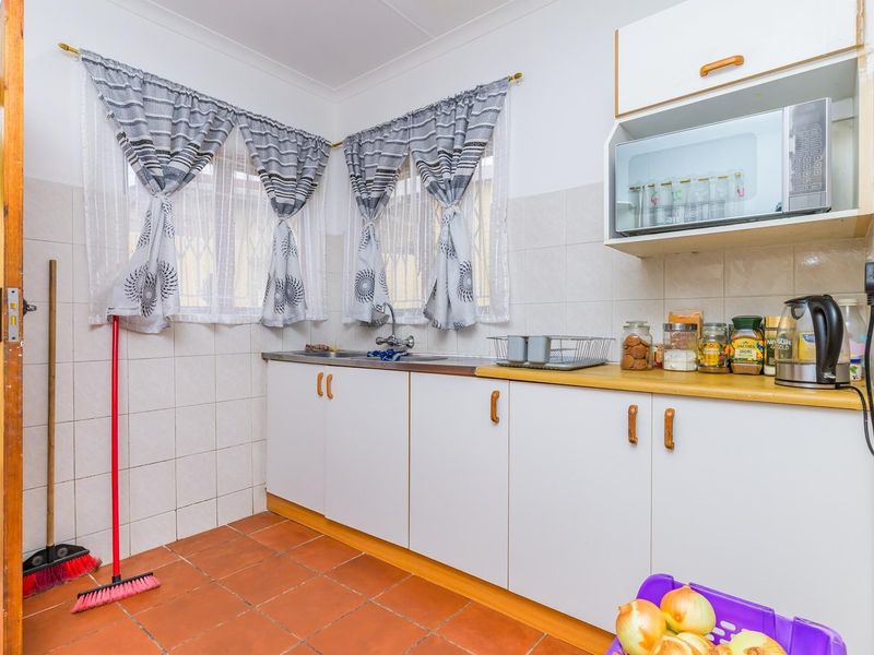 A functional kitchen featuring white cabinetry, a stainless steel sink, tiled splashbacks, and durable terracotta floor tiles with direct access to the exterior.