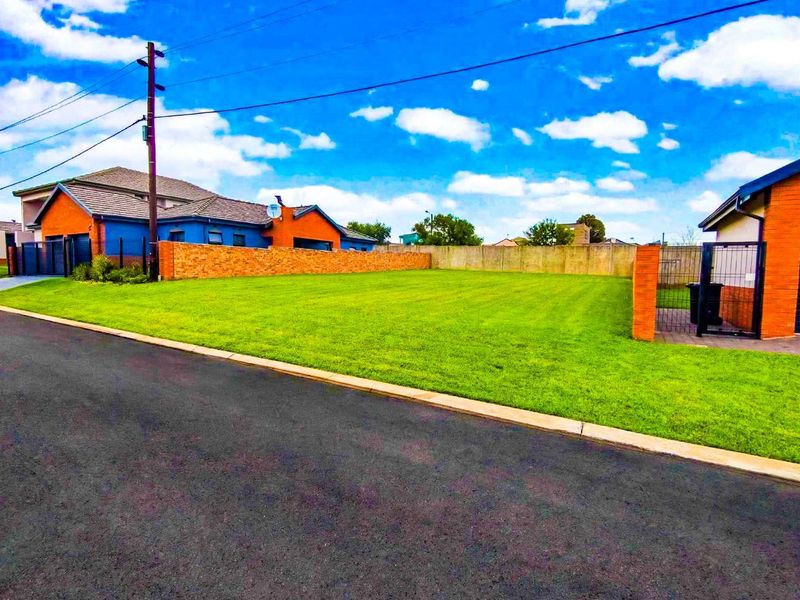 A well-maintained vacant residential plot featuring a manicured lawn, brick boundary walls, and paved street access in a developed neighborhood.