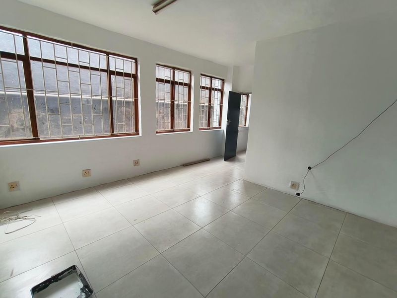 An empty, well-lit office space featuring large windows with security bars, neutral tiled flooring, and functional electrical infrastructure.