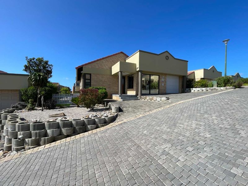 Front exterior view of a single-story brick house featuring a paved driveway, retaining wall landscaping, and a covered entrance with a garage.
