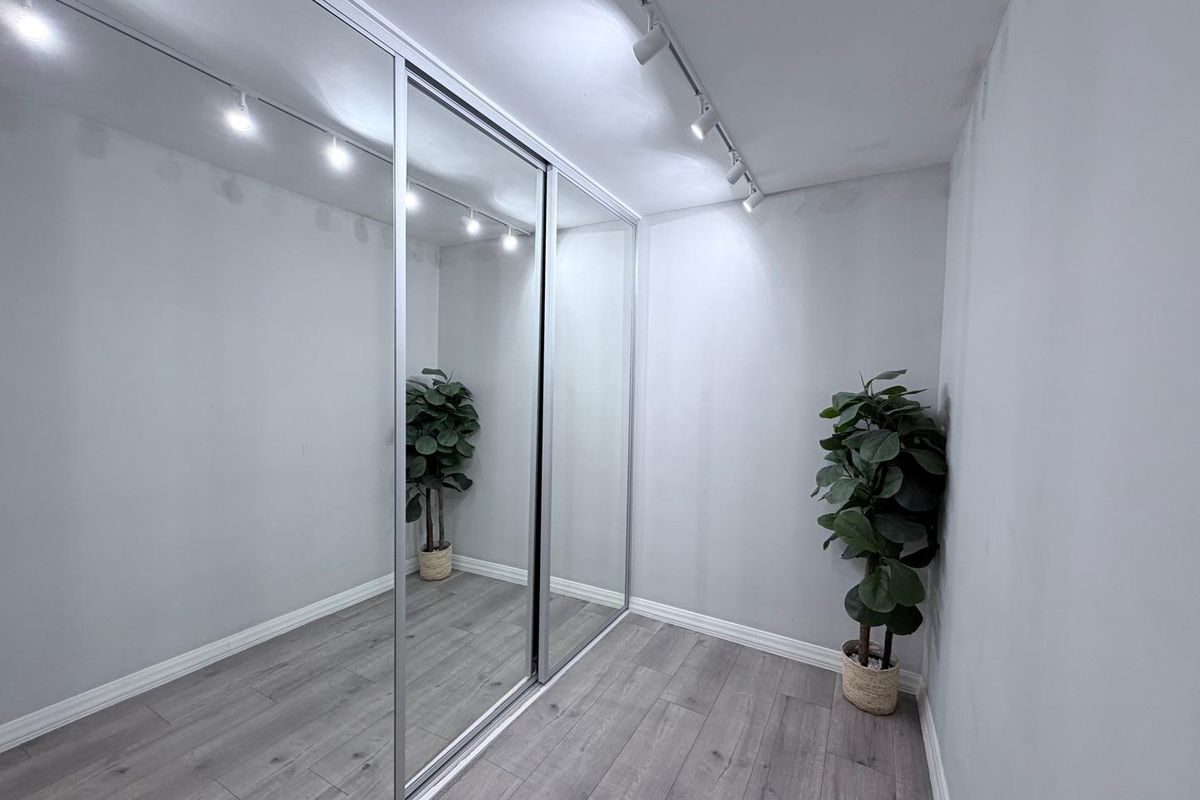 A clean, minimalist bedroom space featuring modern laminate flooring, built-in sliding mirror wardrobe doors, and adjustable track lighting.