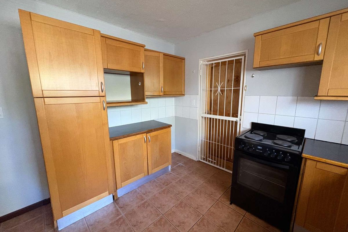 A functional kitchen featuring light wood cabinetry, tiled splashbacks, dark countertops, and a freestanding electric stove, with a security gate leading to the exterior.