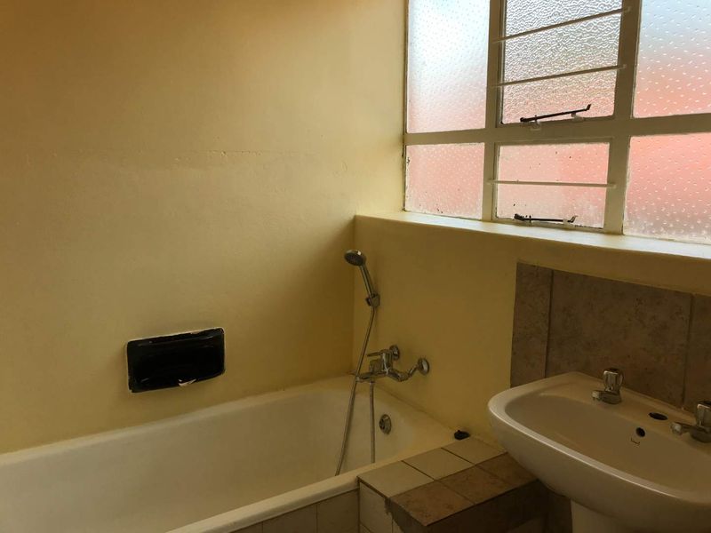 A dated bathroom featuring a bathtub with handheld shower, pedestal basin, and steel-framed window, requiring cosmetic updates to modernize the space.