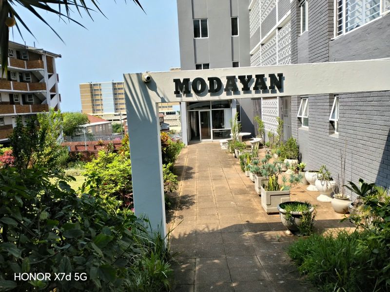 Entrance walkway to the Modayan apartment complex featuring a paved path, landscaped garden beds, and building signage.