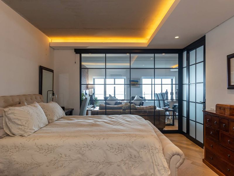 A modern bedroom featuring elegant herringbone timber flooring, recessed ceiling lighting, and a striking black-framed glass partition wall connecting to the living area.