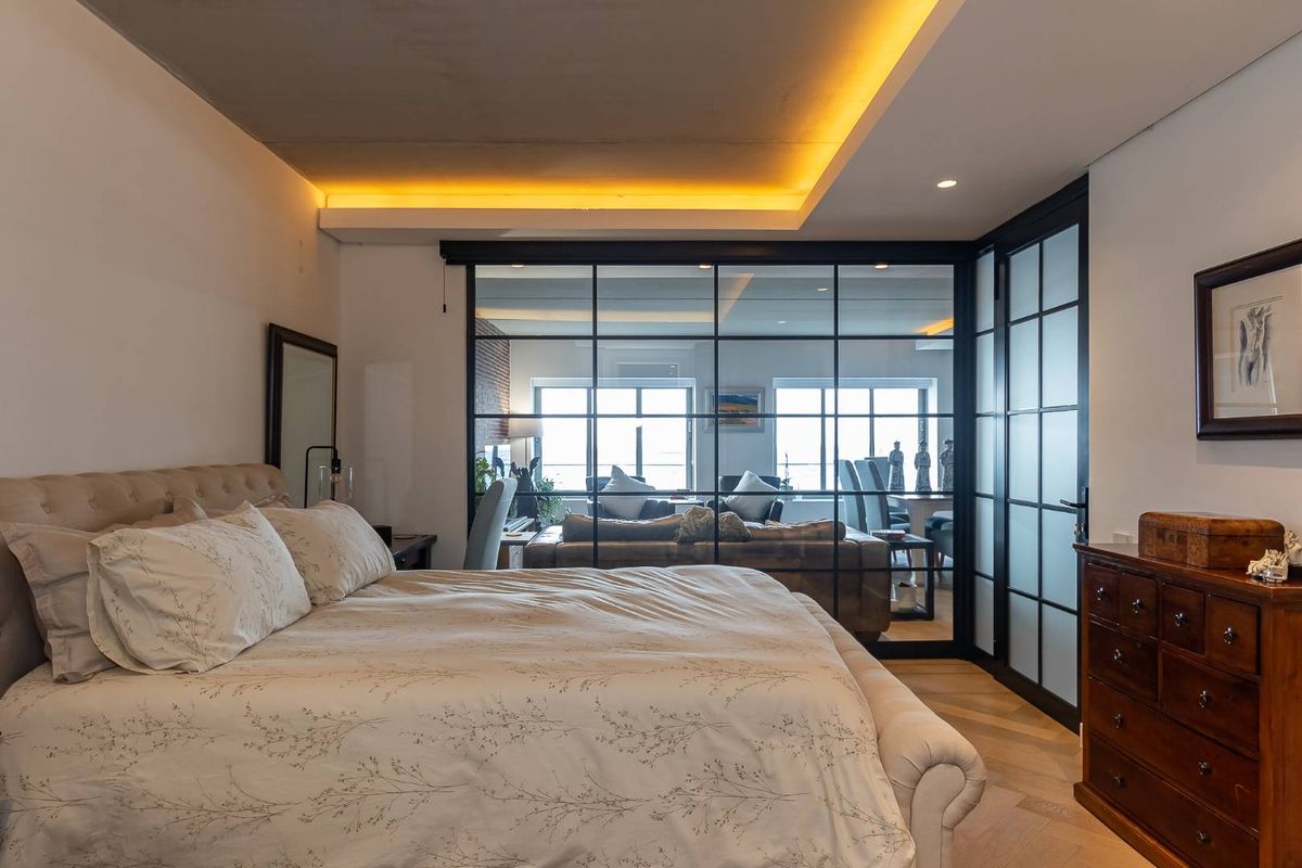 A modern bedroom featuring elegant herringbone timber flooring, recessed ceiling lighting, and a striking black-framed glass partition wall connecting to the living area.