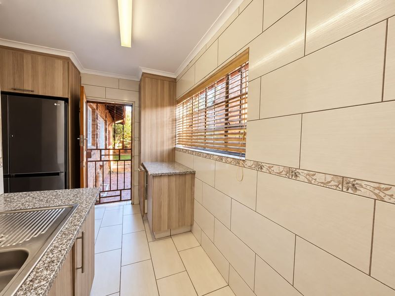 A clean and functional scullery featuring modern wood-look cabinetry, granite countertops, tiled walls, and direct access to the exterior through a security gate.