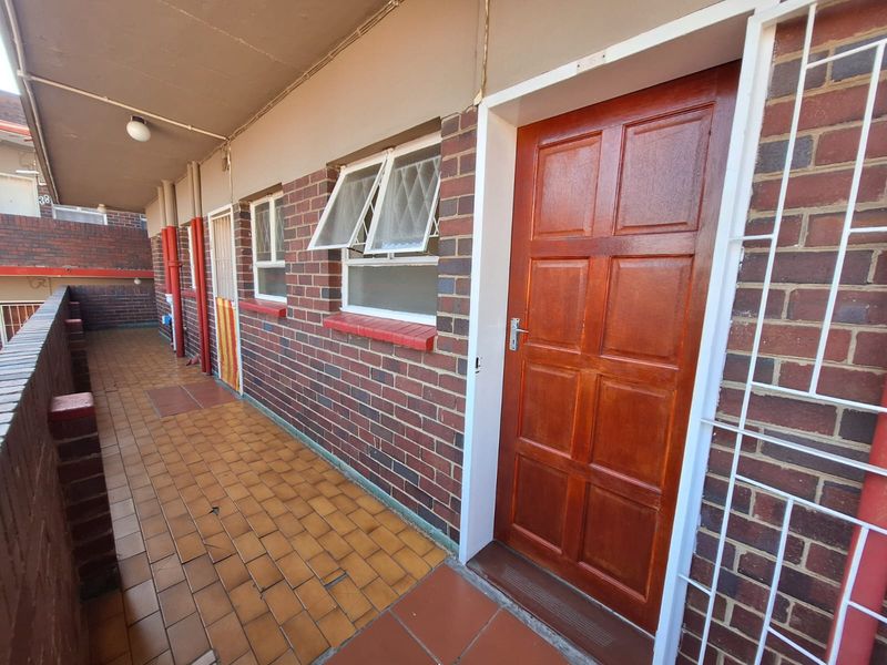 An exterior balcony area featuring brick walls, tiled flooring, and a secure wooden entrance door with security bars.
