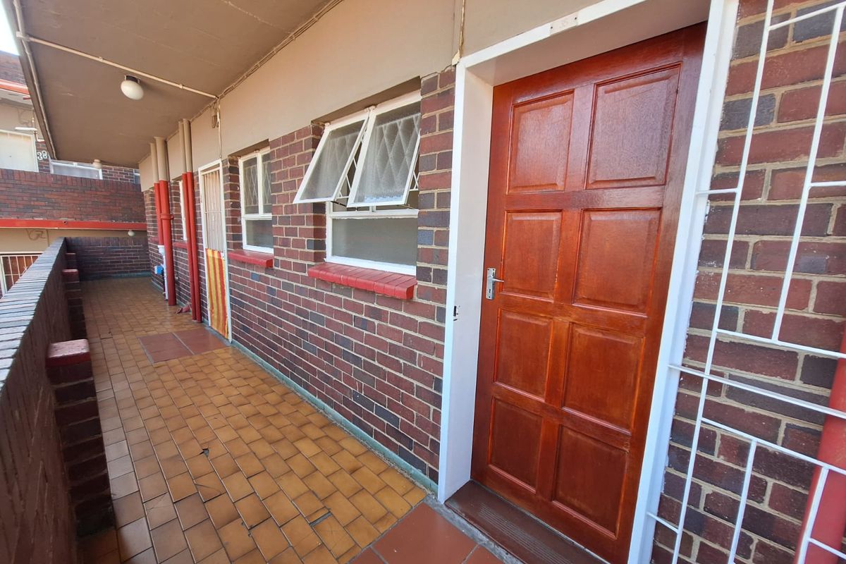 An exterior balcony area featuring brick walls, tiled flooring, and a secure wooden entrance door with security bars.