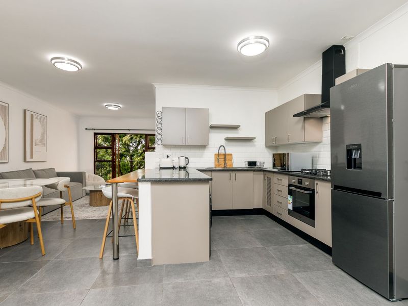 Modern open-plan kitchen and living area featuring sleek cabinetry, stone countertops, integrated appliances, and large format floor tiles leading to a balcony.