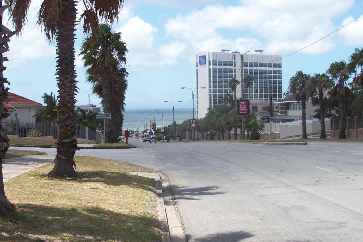 A street view showing the surrounding neighbourhood approach, featuring palm-lined roads and a view towards the coast and nearby high-rise buildings.