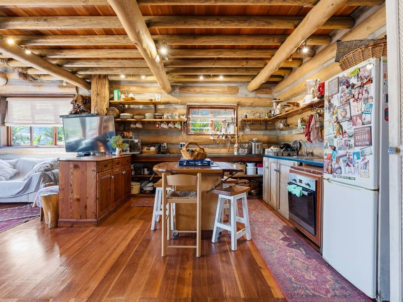 Rustic open-plan living and kitchen area featuring exposed log walls, timber beam ceilings, hardwood floors, and integrated kitchen cabinetry with built-in oven.