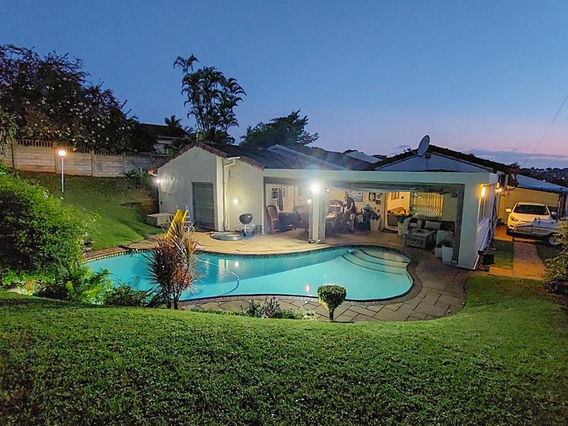 Rear view of the property featuring a swimming pool, paved patio area, and landscaped garden at dusk.