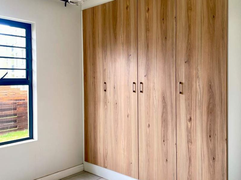 A bright, empty bedroom featuring modern wood-finish built-in cupboards, neutral tiled flooring, and a contemporary pendant light fixture.