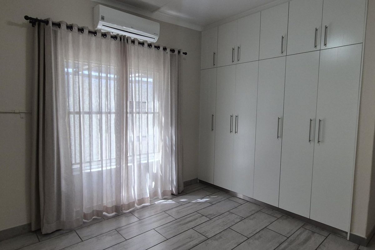 A bright, empty bedroom featuring extensive built-in cupboards, tiled flooring, and a wall-mounted air conditioning unit.