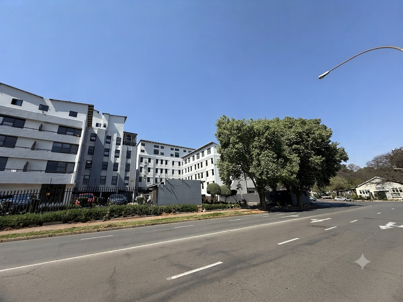 A street-level view of the apartment complex facade, showing the building exterior, perimeter security fencing, and adjacent road infrastructure.