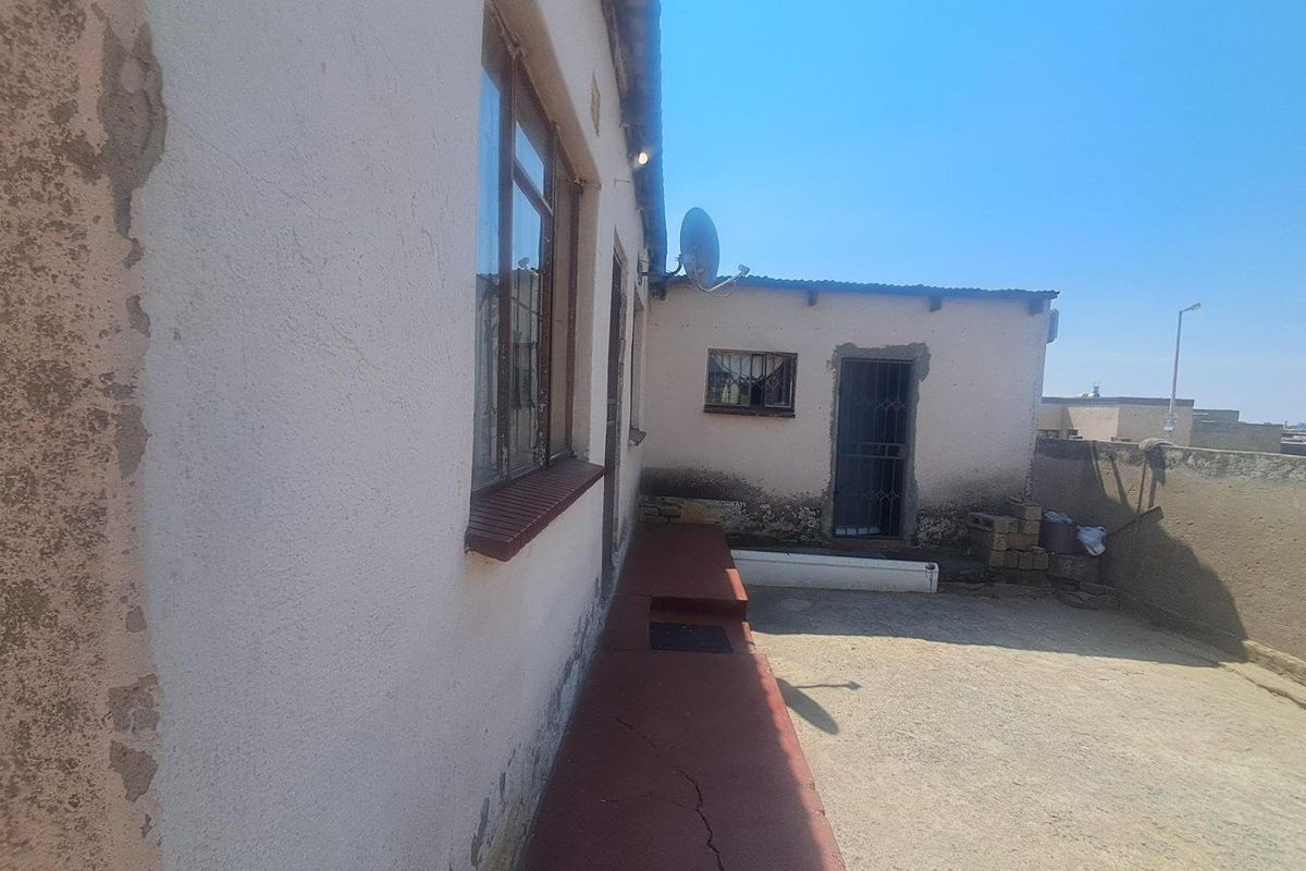 Rear exterior view of the property showing a concrete patio area, weathered wall finishes, and security features on windows and doors.