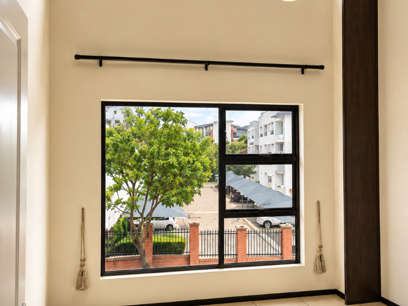 A bright, empty living area featuring double-volume windows, tiled flooring, and modern architectural details, offering a pleasant view of the complex grounds.