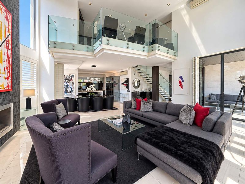 Spacious open-plan living area featuring a double-volume ceiling, mezzanine level with glass balustrade, tiled floors, and seamless flow to an outdoor patio.