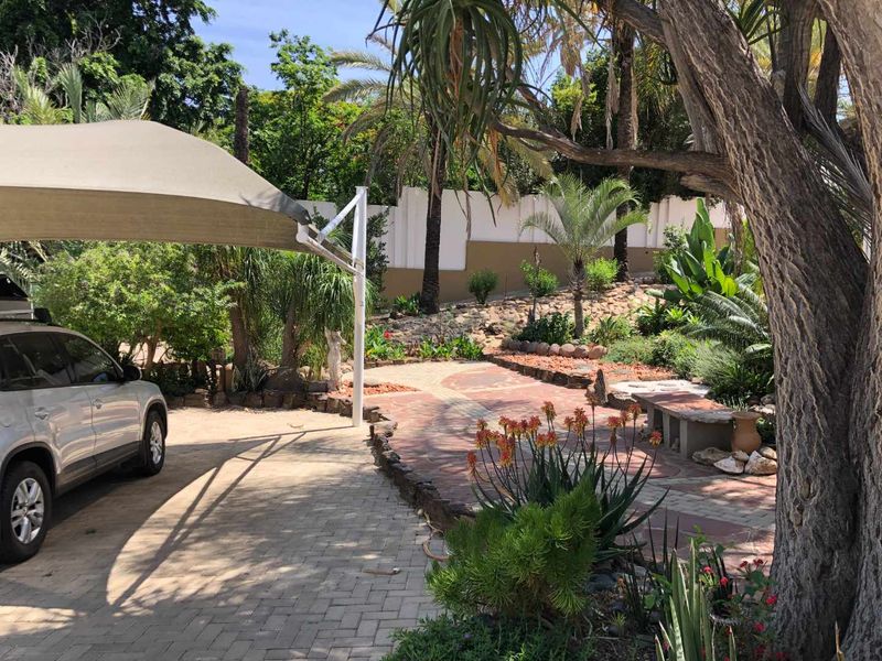 A well-maintained front exterior area featuring a paved driveway with a shade sail, stone retaining walls, and established garden landscaping.
