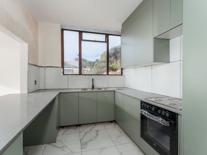 A modern kitchen featuring sage green cabinetry, stone countertops, and built-in appliances, currently undergoing final finishing touches to the walls and tiling.