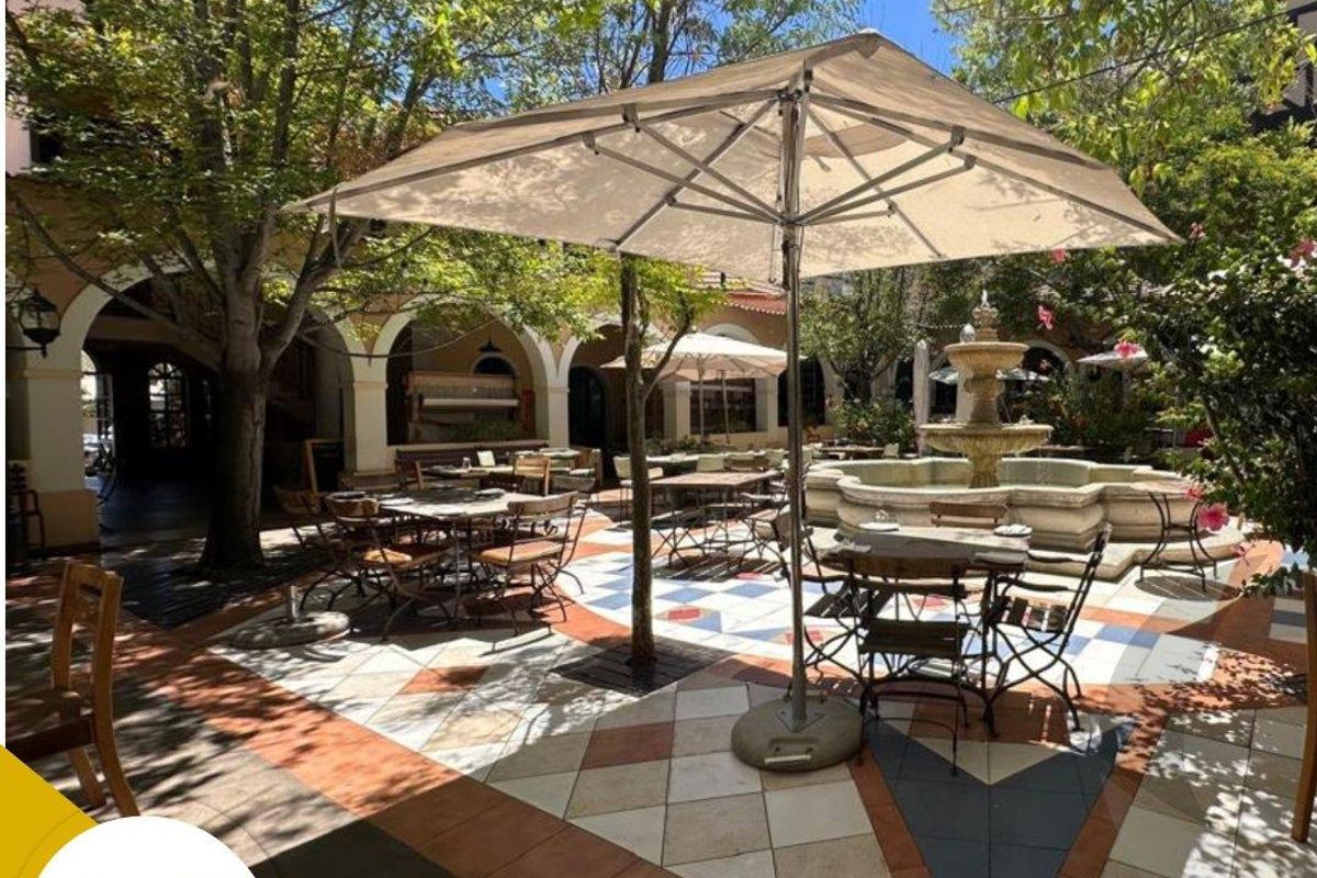 A charming outdoor courtyard featuring a central stone fountain, decorative patterned tile flooring, and an arched colonnade providing a pleasant shaded seating area.