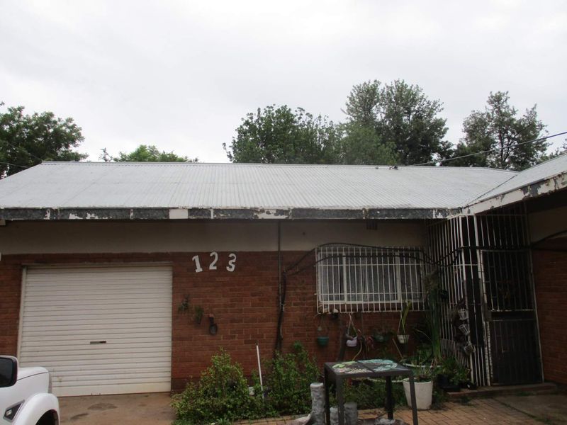 Front exterior view of a single-story brick house featuring a corrugated metal roof, roller garage door, and security bars on windows.