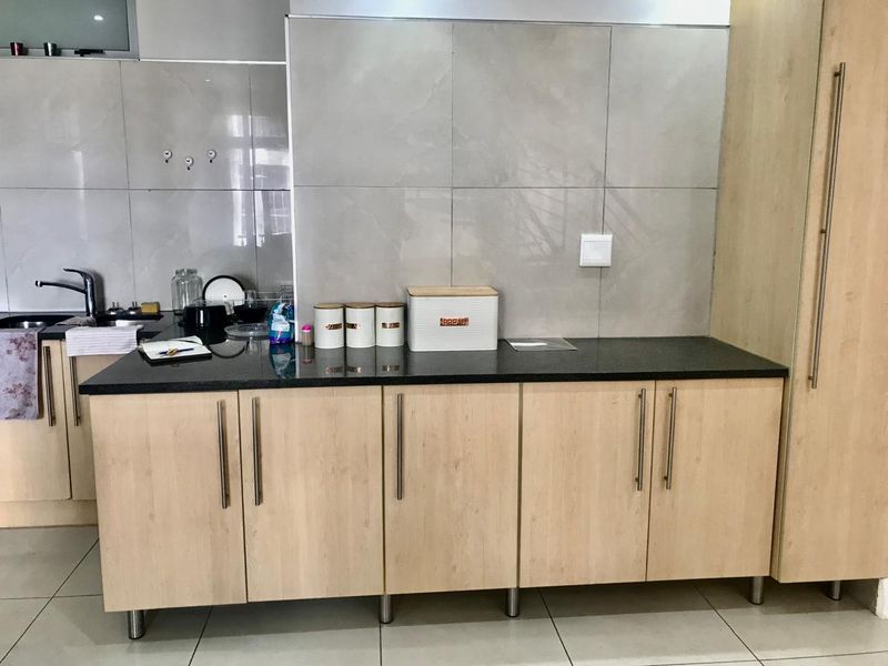 A functional kitchen area featuring light wood cabinetry, dark granite countertops, a stainless steel sink, and modern large-format wall tiling.