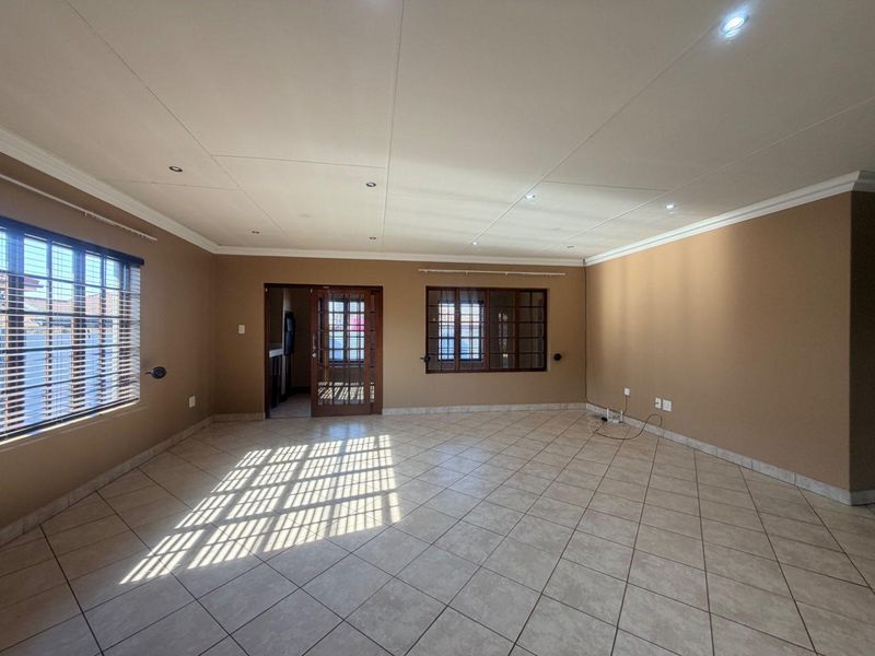 A spacious, empty living room featuring neutral-toned walls, tiled flooring, recessed downlighting, and timber-framed windows and doors.