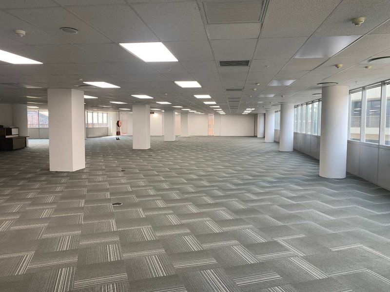 A spacious, open-plan office area featuring a suspended ceiling, recessed lighting, carpet tiles, and large windows providing ample natural light.