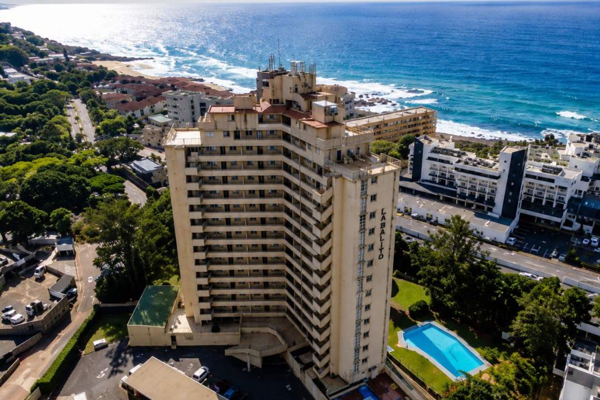 Aerial view of a high-rise apartment complex featuring a swimming pool, landscaped grounds, and covered parking, situated near the coastline.