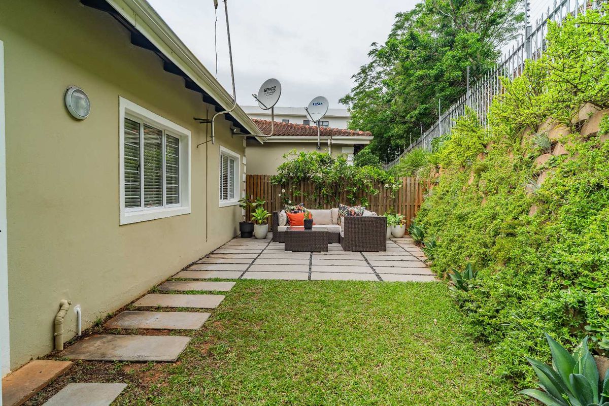 A private rear garden featuring a paved patio area, manicured lawn, and a terraced retaining wall with established greenery.