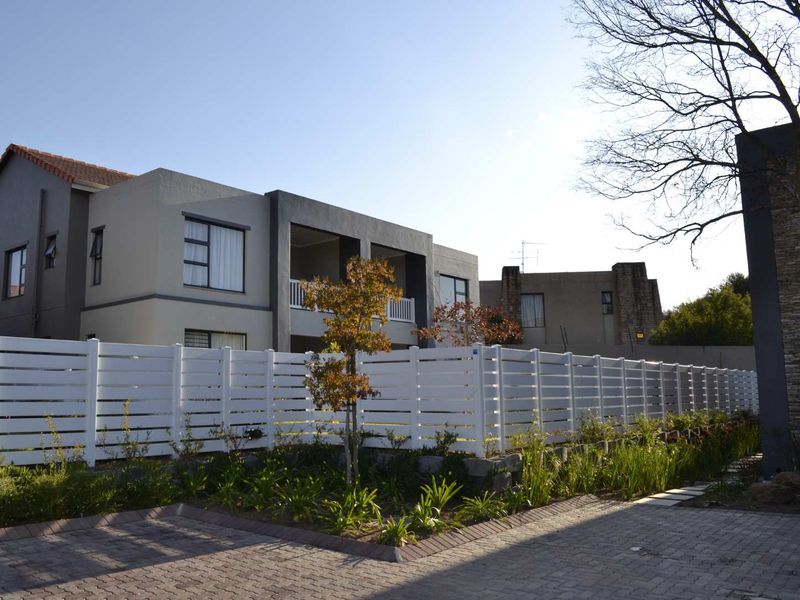 Exterior view of a modern apartment complex featuring a paved driveway, white perimeter fencing, and landscaped garden areas.