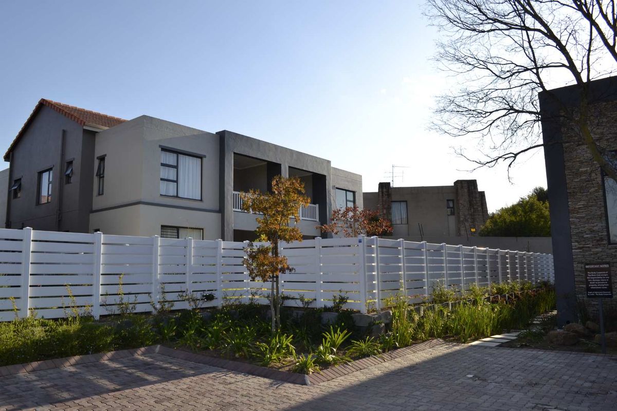 Exterior view of a modern apartment complex featuring a paved driveway, white perimeter fencing, and landscaped garden areas.