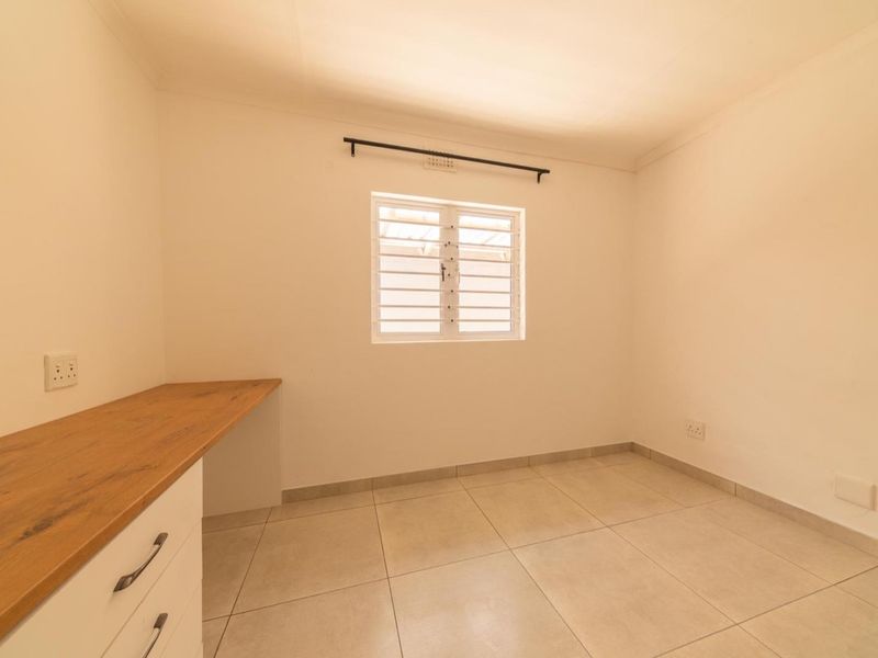 A clean, empty bedroom featuring neutral tiled flooring, a built-in wooden desk, and a louvered window with a curtain rod.