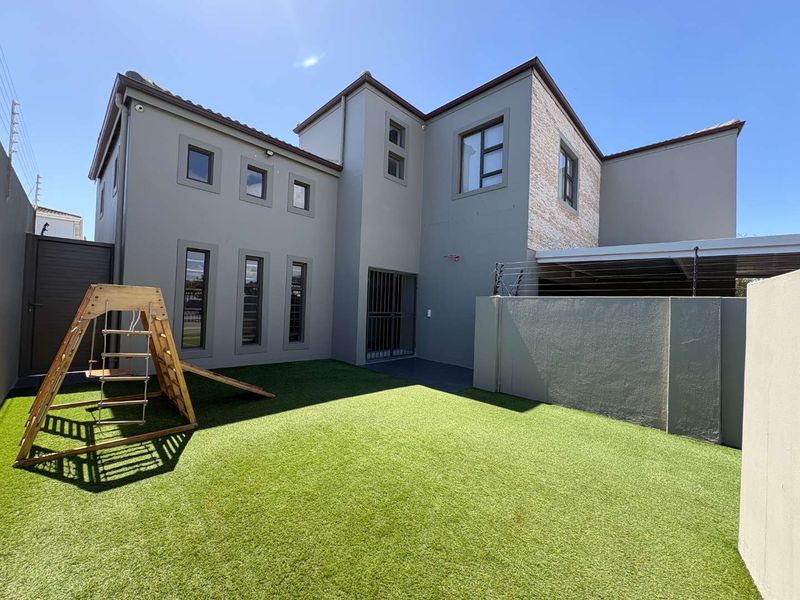 A well-maintained rear garden featuring artificial turf, a secure perimeter wall with electric fencing, and a modern double-story building facade.