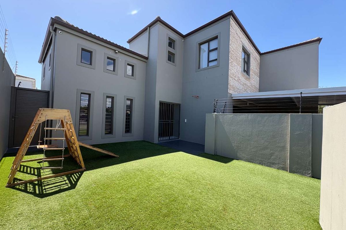A well-maintained rear garden featuring artificial turf, a secure perimeter wall with electric fencing, and a modern double-story building facade.