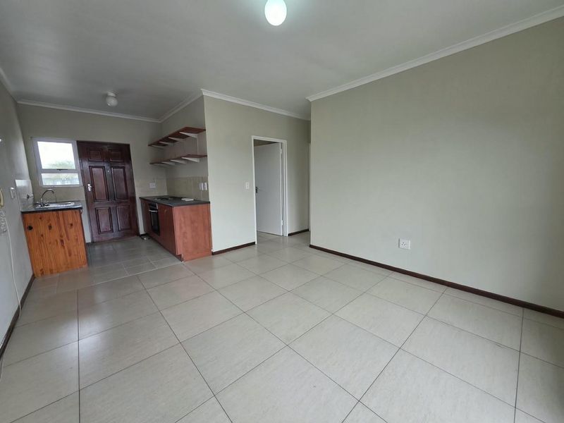 An open-plan living and kitchen area featuring tiled flooring, built-in cabinetry with an integrated oven and hob, and a wooden entrance door.