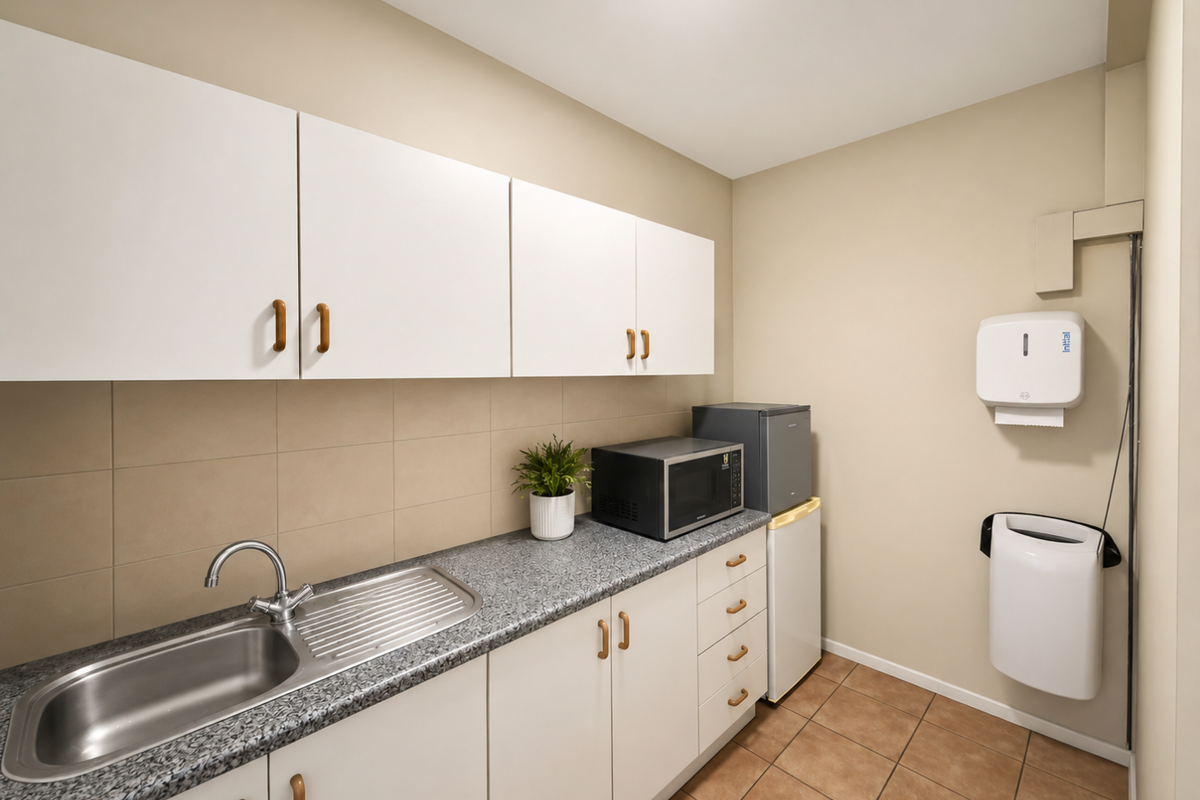A functional kitchenette area featuring white cabinetry, a stainless steel sink, granite-look laminate countertops, and wall-mounted utility dispensers.
